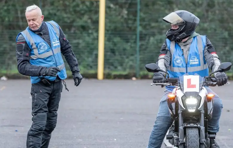 An instructor in a blue vest walks across the training ground while a learner in a black helmet sits on a motorbike, looking towards them.