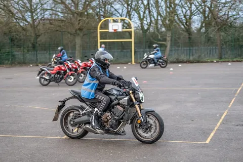 A learner in a blue student vest rides a dark motorbike across the training ground, with a row of red bikes parked behind.