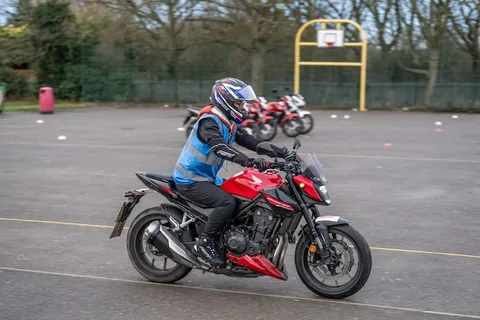 A learner in a blue vest and blue-and-red helmet leans a red and black motorbike into a turn on the practice area.