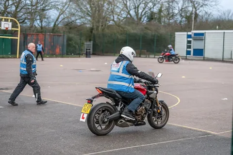 A learner in a white helmet turns a dark motorbike while an instructor in a blue vest walks across the training ground behind.