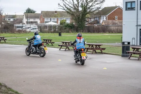 Two learners ride at a distance across a grey practice area, with picnic benches and houses behind them.