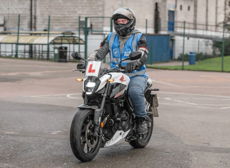 A learner in a blue student vest and grey helmet rides a white and red motorbike with an L plate across the training ground.