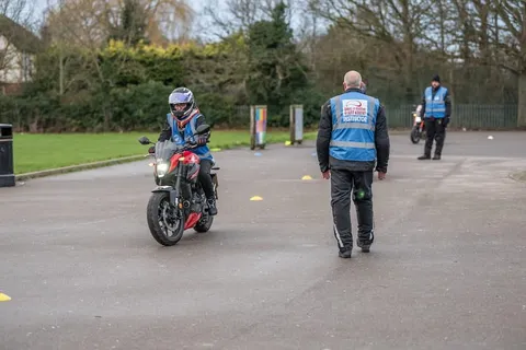 A learner waits on a bike at the left while an instructor in a blue vest walks across the practice area and a second instructor looks on.