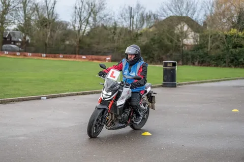 A learner in a grey helmet rides a white and red motorbike around a yellow cone, with a wide green field behind.