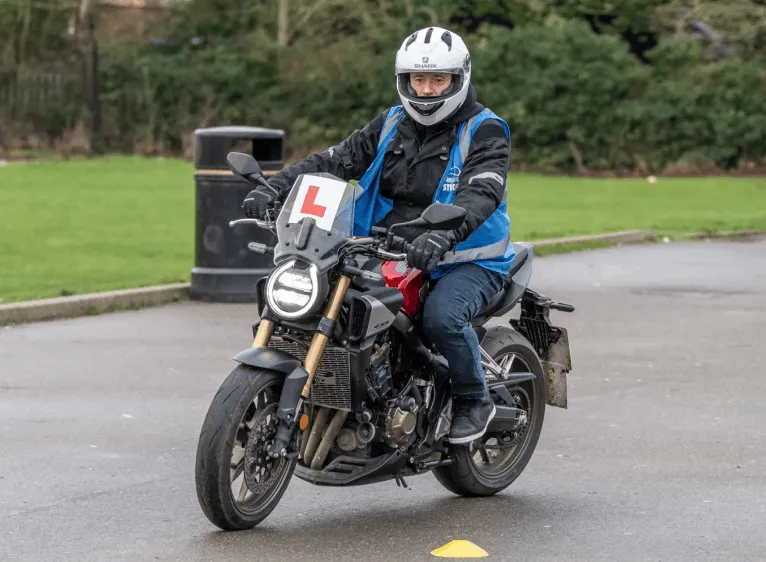 A learner in a white helmet and blue student vest rides a dark motorbike with an L plate past a bin, with a green field behind.