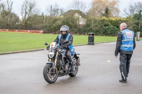 A learner in a white helmet rides a dark motorbike on wet tarmac while an instructor in a blue vest stands to one side.