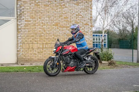 A learner in a blue vest and blue helmet rides a red motorbike past a yellow-brick building.