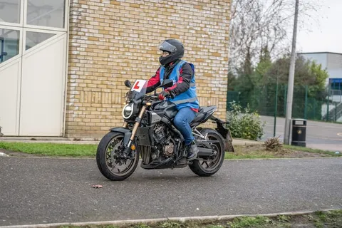 A learner in a black helmet and blue vest rides a dark motorbike with an L plate in front of a yellow-brick school building.