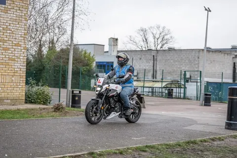 A learner in a blue vest rides a white motorbike along a path, with a brick wall, a street lamp and school buildings behind.