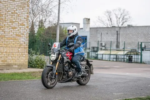A learner in a white helmet rides a dark motorbike with a red frame towards the camera, beside a brick wall.