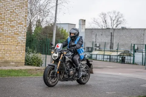 A learner in a black helmet rides a dark motorbike with an L plate near a brick wall and school buildings.