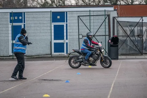 An instructor in a blue vest gestures from the edge of the practice area while a learner rides a dark bike past blue and yellow cones.