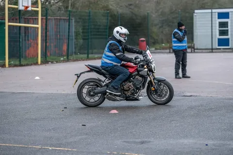 A learner in a white helmet rides a dark motorbike around a pink cone while an instructor and another person watch.