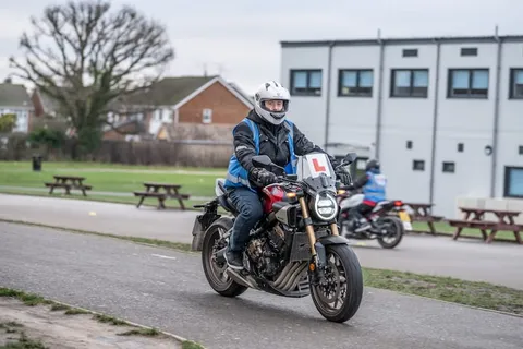 A learner in a white helmet rides a dark motorbike towards the camera on a curved path, with picnic tables and a white building behind.