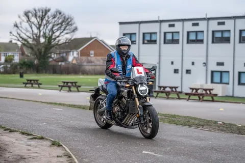 A learner in a black helmet and red jacket rides a dark motorbike along a curved path in front of a white two-storey building.