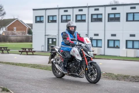 A learner in a white helmet rides a red and white motorbike along the practice path, with a white building and houses behind.
