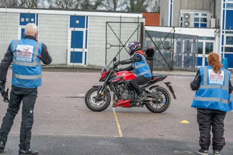 Two instructors in blue vests stand either side of the practice area, watching a learner ride a red motorbike between them.