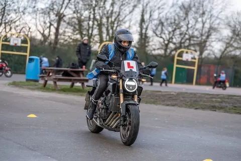 A learner rides a dark motorbike straight towards the camera past yellow cones, with basketball hoops and other learners behind.