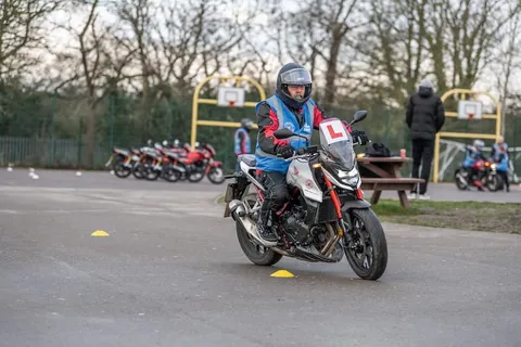 A learner in a red and blue jacket rides a white motorbike towards the camera, with a row of parked bikes and basketball hoops behind.