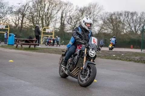 A learner in a white helmet rides a dark motorbike towards the camera on an overcast day, with parked bikes and picnic tables behind.