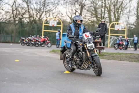 A learner in a grey helmet rides a dark motorbike past a yellow cone, with a line of parked red bikes and an instructor watching.
