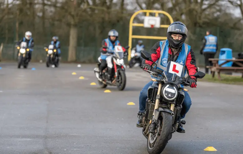 A learner in a blue student vest rides a motorbike towards the camera, with more learners riding behind.