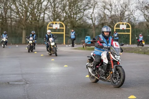 A learner rides a white and red motorbike along a line of yellow cones, with more riders coming up behind.