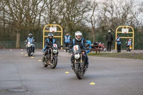 Two learners ride towards the camera along a lane marked with yellow cones, with more riders and instructors behind them.