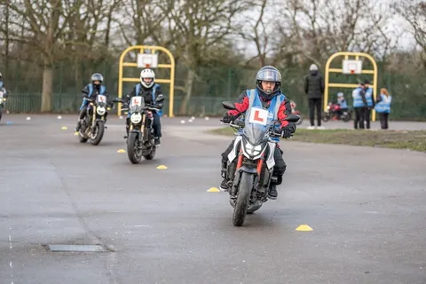 Three learners follow each other along a cone lane, the nearest in a white helmet on a white and red motorbike.