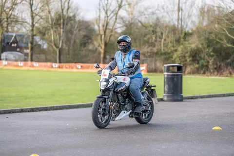 A learner in a black helmet rides a white and grey motorbike past a bin, with a wide green field behind.