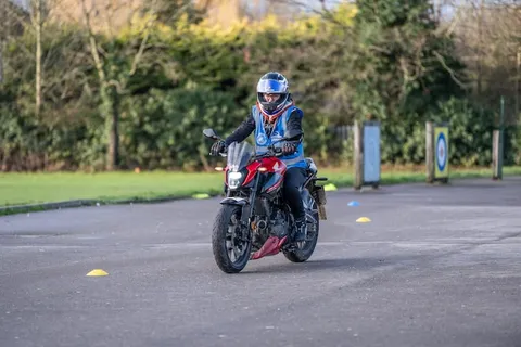 A learner in a blue helmet rides a red motorbike past yellow cones, with hedges and target boards behind.