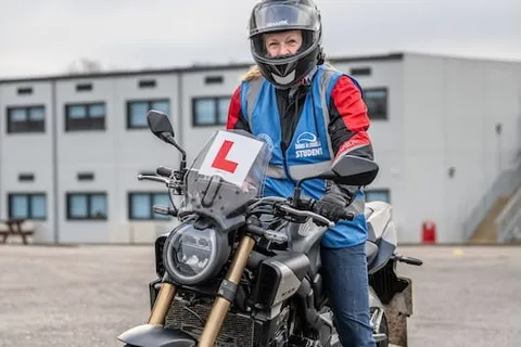 A learner in a black helmet sits on a dark motorbike with an L plate in front of a white two-storey building.