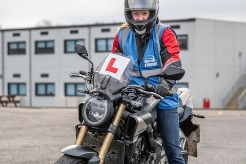 A learner in a grey helmet sits on a dark motorbike with an L plate, with a white two-storey building behind.