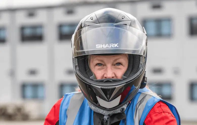 A close-up of a learner in a full-face helmet and a blue hi-vis vest, looking straight at the camera.