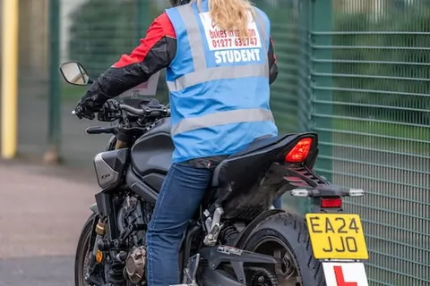 A learner with a long ponytail, seen from behind in a blue student vest, sits on a dark motorbike beside a green fence.