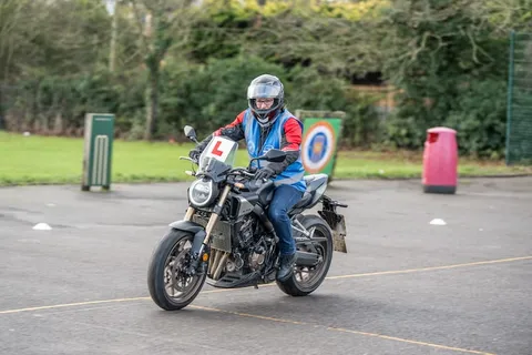 A learner in a grey helmet rides a dark motorbike across the practice area, with hedges, a bin and a target board behind.