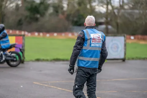 An instructor in a blue vest, seen from behind, stands watching over the practice area with a target board in the distance.