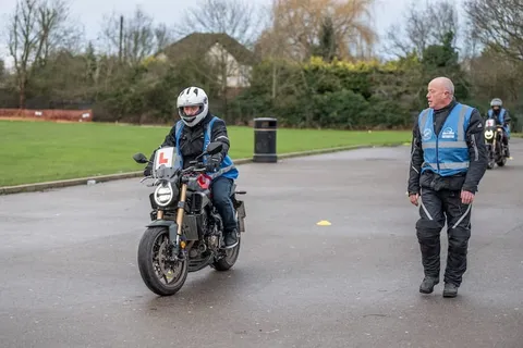 A learner on a dark motorbike rides towards an instructor in a blue vest standing on the wet tarmac.