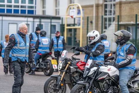 An instructor in a blue vest talks to learners waiting on their bikes, with other learners and instructors behind them.
