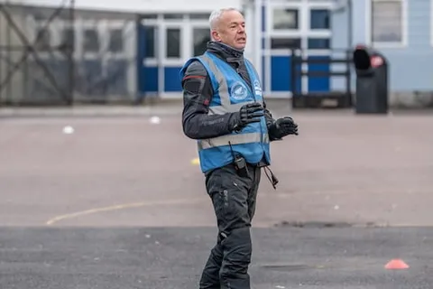 An instructor in a blue vest stands alone on the practice area, gesturing with one hand.