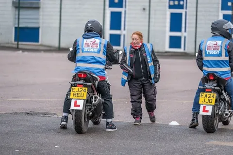 An instructor in a blue vest stands between two learners on parked motorbikes, briefing them before an exercise.