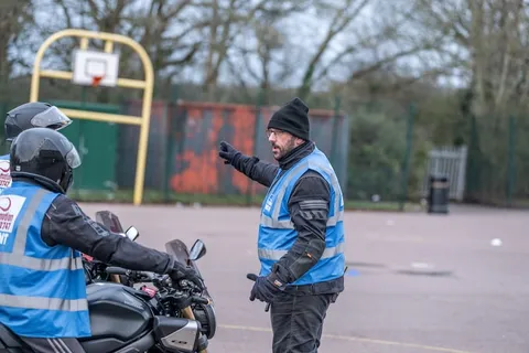An instructor in a beanie and blue vest points while explaining an exercise to a learner on a dark motorbike.