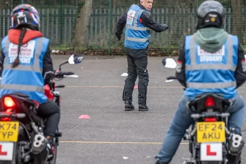 Two learners on motorbikes with yellow number plates wait side by side while an instructor stands in the distance.