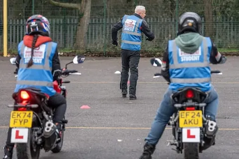 Two learners on motorbikes wait side by side, facing away from the camera, as an instructor stands ahead of them.