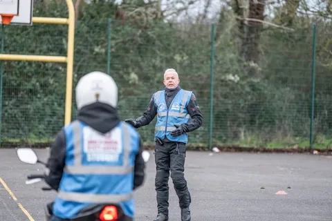 An instructor in a blue vest gestures while speaking to a learner in a white helmet, who is seen from behind.