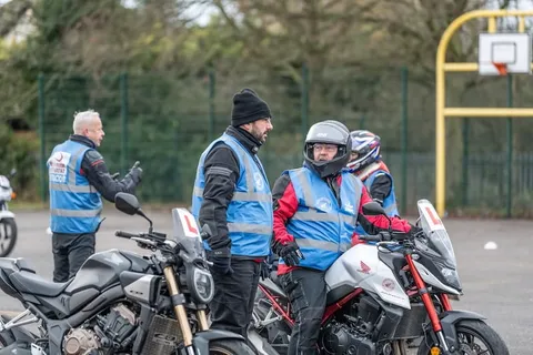Two instructors talk to a learner on a white and red motorbike, with a dark motorbike in the foreground.