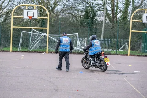 An instructor in a blue vest watches a learner on a dark motorbike ride away across the practice area.