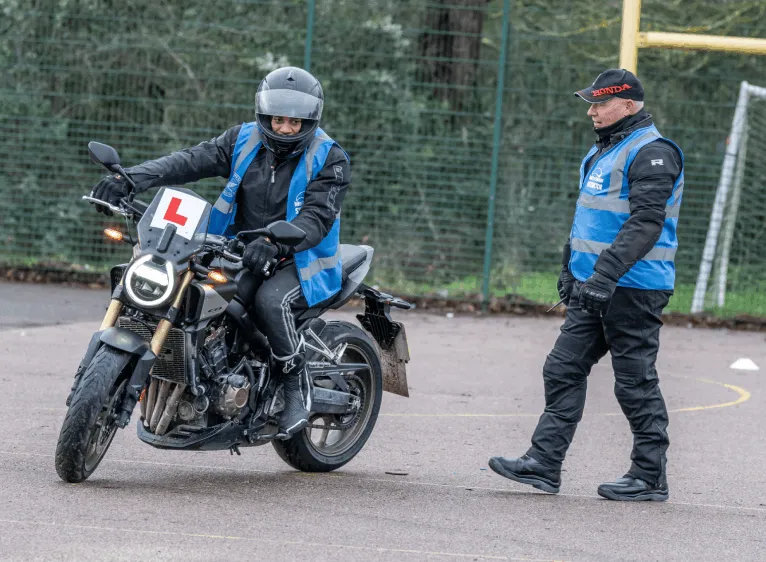A learner in a black helmet and blue vest stops a motorbike as an instructor in a Honda cap walks towards them.