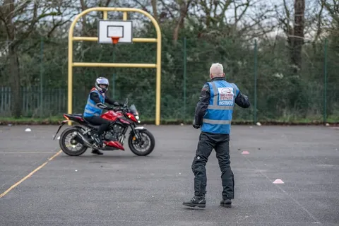A learner on a red motorbike rides across the practice area while an instructor in a blue vest watches with their back to the camera.