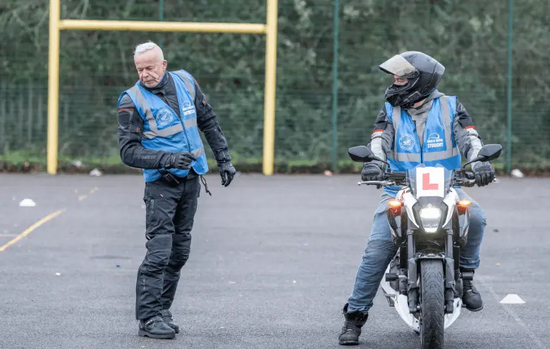 An instructor in a blue vest walks across the training ground while a learner in a black helmet sits on a motorbike, looking towards them.